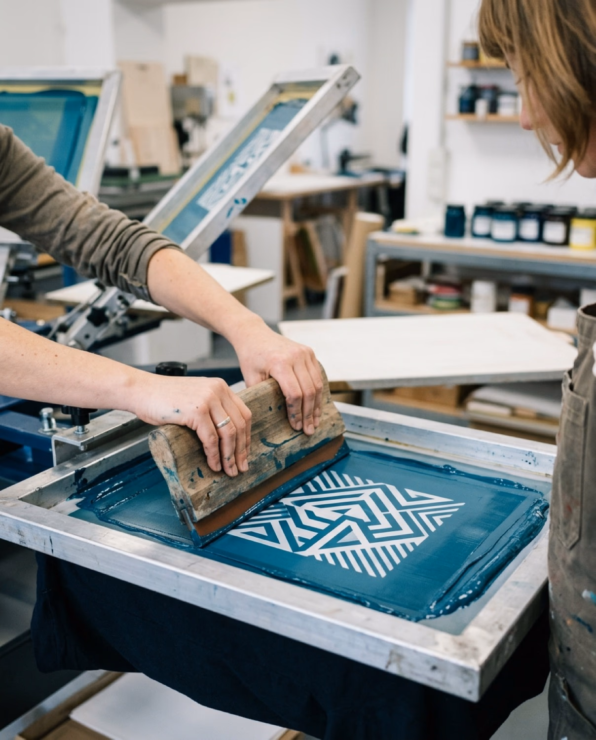 Druckerin presst petrol-blaue Siebdruckfarbe mit Squeegee durch einen Druckrahmen in der Sportatex-Werkstatt – geometrisches Vereinslogo wird sichtbar